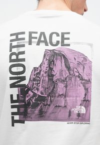 Λευκό μπλουζάκι με μεγάλο μοβ γραφικό βουνού και δέντρων, με την επιγραφή "THE NORTH FACE" από πάνω και "NEVER STOP EXPLORING" από κάτω.