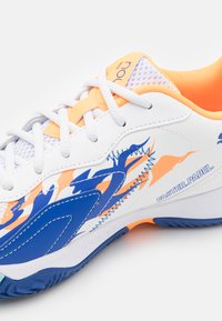 Chaussure de sport blanche avec des accents bleu et orange, présentant une surface texturée, un panneau en maille et une semelle courbée pour l'adhérence et le soutien.