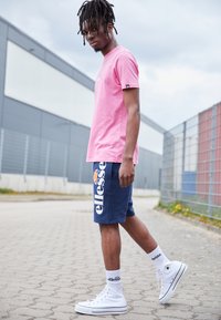 Rosa Kurzarm-T-Shirt, marineblaue Shorts mit "ellesse" Logo, weiße High-Top-Sneaker und weiße Socken. Lässiger Streetwear-Stil.