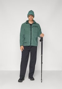 Giacca in pile verde con zip e due tasche, abbinata a pantaloni neri e scarpe da trekking. Include un berretto abbinato e un bastone da trekking.