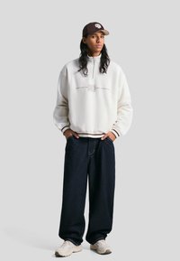 Bela, zadržeča jakna s kapuco in zadrgo z napisom "BOLD" ter rjavimi detajli. Ohlapni temno modri široki trije hlače in bež superge. Kratka kapa s ukrivljenim robom.