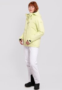 Hellgrüne, wasserdichte Jacke mit Kapuze, mit Reißverschluss verschließbaren Taschen und verstellbaren Bündchen, kombiniert mit weißen Hosen und schwarzen Stiefeln.