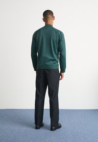 Mörkgrön jacka med ribbad krage och muddar, ihop med svarta byxor och skor. Slät tygkvalitet med en figurnära design och inga synliga mönster.