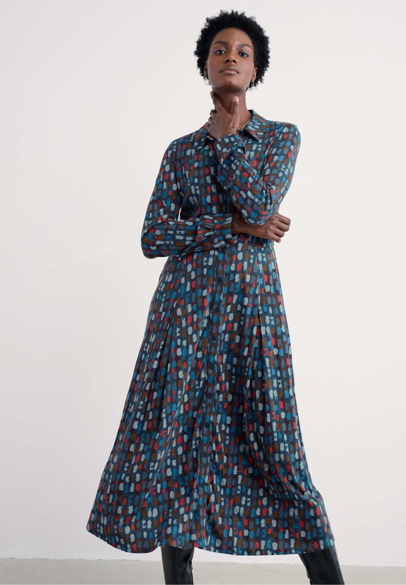 Nainen, jolla on lyhyet kiharat hiukset, seisoo käsi kaulalla ja hänellä on pitkä nappimekko, jossa on sinisiä, punaisia ja harmaita abstrakteja kuvioita.