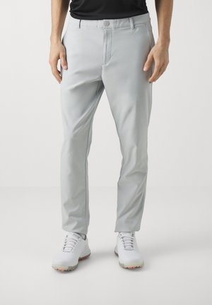 Homme portant un pantalon slim gris clair et des baskets blanches lacées, debout devant un fond uni.