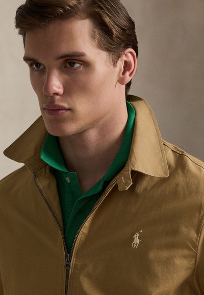 Καφέ μπουφάν με φερμουάρ πάνω από ένα πράσινο πόλο πουκάμισο. Το μπουφάν διαθέτει ψηλό γιακά και μικρό λογότυπο στο αριστερό στήθος. Ομαλή υφή υφάσματος.
