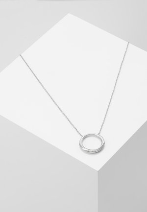 Collier en argent avec un pendentif circulaire présentant un design lisse et de petites pierres incrustées, exposé sur une surface géométrique blanche.