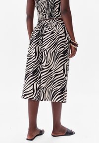 Zwart en beige jurk met zebra-patroon, met een nauwsluitende smockbovenkant en een losse rok. Model draagt platte zwarte sandalen en gestapelde armbanden.