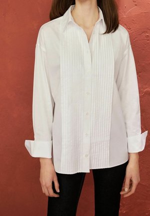 Chemise blanche à manches longues en tissu lisse, avec des plis verticaux sur le devant et une fermeture à boutons. Les poignets sont retroussés.