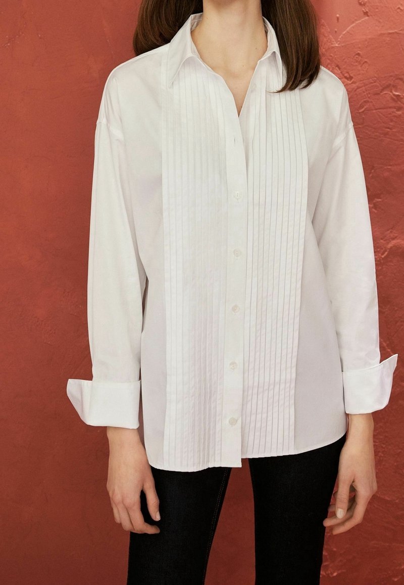 Chemise blanche à manches longues en tissu lisse, avec des plis verticaux sur le devant et une fermeture à boutons. Les poignets sont retroussés.