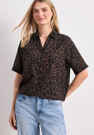 Femme portant un chemisier à manches courtes imprimé léopard marron et noir et un jean bleu clair, debout avec une main dans le dos.