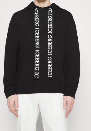 Musta huppari, jossa on suuri valkoinen "ICEBERG"-teksti kiristysnauhoissa, päällä henkilö valkoisissa housuissa vaaleaa taustaa vasten.