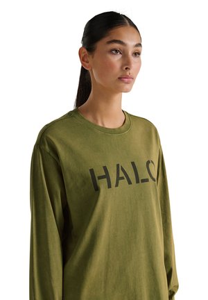 Jonge vrouw met donker haar in een paardenstaart, gekleed in een olijfgroene, lange mouwen shirt met "HALO" in het zwart gedrukt op de voorkant.