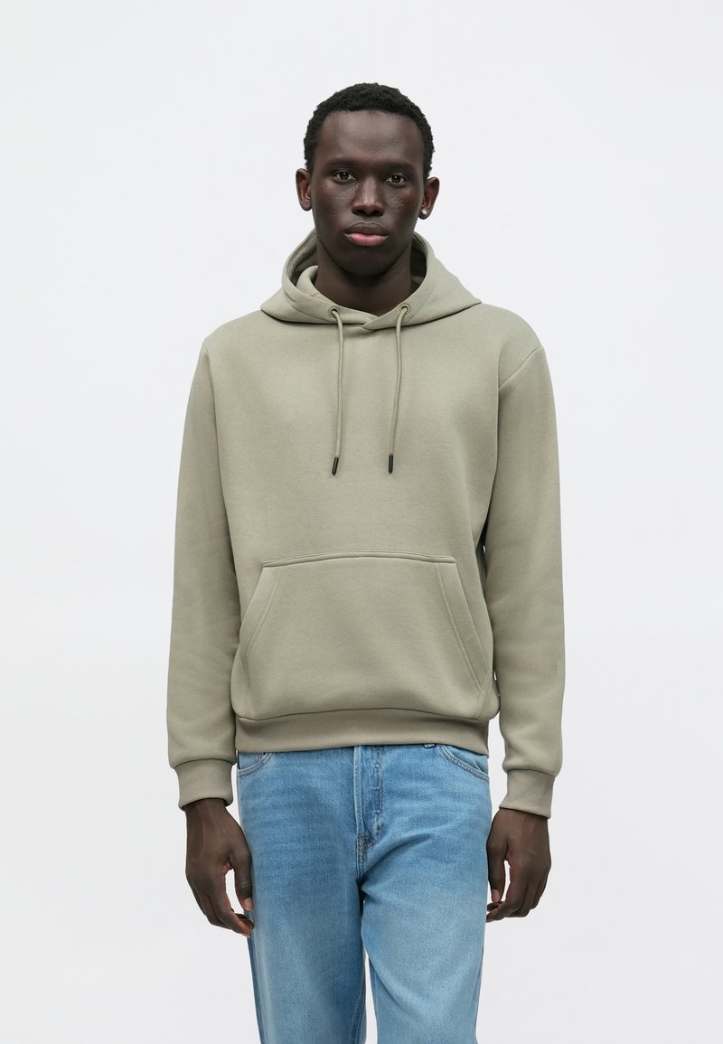 Hettegenser i lys olivengrønn, med frontlomme, snorer og ribbestrikkede mansjetter. Kombinert med lyseblå jeans.