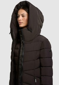 Giacca nera imbottita con colletto alto e cappuccio oversize. Caratteristiche includono una chiusura con zip frontale e tessuto testurizzato con pannelli cuciti.