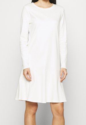 Femme portant une robe blanche à manches longues, longueur genou, avec un col rond et un ourlet évasé, debout devant un fond uni.