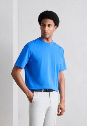 Άνδρας με αφρο χτένισμα που φοράει φωτεινό μπλε μπλουζάκι και ανοιχτό γκρι παντελόνι, στέκεται με το ένα χέρι στην τσέπη μπροστά σε απλό φόντο.