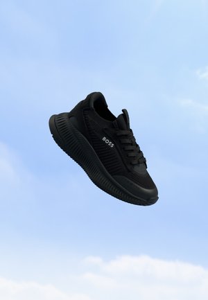 Sort BOSS-sneaker med struktureret sål og snørebånd, der svæver mod en klar blå himmelbaggrund.