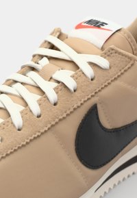 Bežiniai zomšiniai ir audiniai sportbačiai su baltais raišteliais, juodu Nike logotipu ir raudonu bei baltu žymekliu ant liežuvio. Klasikinis dizainas su tekstūriškais akcentais.