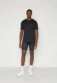 Schwarzes Kurzarm-T-Shirt mit Logo, kombiniert mit schwarzen Sportshorts. Das Modell trägt weiße Socken und Turnschuhe und steht vor einem schlichten Hintergrund.