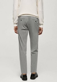 Homme portant un pantalon ajusté gris clair, un pull crème et des chaussures habillées noires, debout devant un fond clair uni.