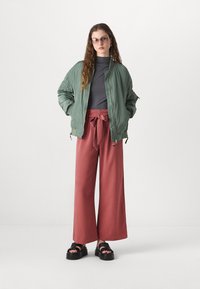 Green bomber jacket with a nylon texture, high-neck gray top, wide-leg reddish-pink trousers tied at the waist, and chunky black platform sandals.