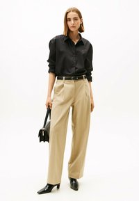 Zwarte knopshirt met opgerolde mouwen, beige high-waisted broek, zwarte riem en zwarte puntige haklaarzen. Houdt een zwarte handtas vast.