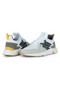 Zapatillas deportivas en blanco y gris con parte superior de malla, detalles en ante, un elemento de diseño negro y una pestaña en el talón amarilla con suela de goma.