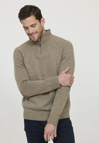 Pull en laine beige avec un col zippé mi-hauteur, des poignets et un ourlet côtelés, ainsi qu'une texture subtile. Présente un petit logo sur la manche.