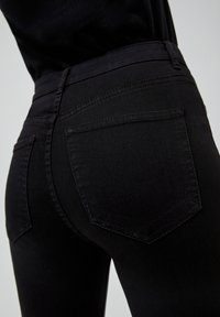 Jeans de denim negro con un diseño ajustado, talle alto y dos bolsillos traseros con un patrón de costura visible. Textura lisa en toda la prenda.