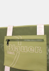 Borsa a rete verde con tasca frontale con zip, logo stampato in bianco e manici rosa pallido. Il materiale testurizzato offre un aspetto moderno.
