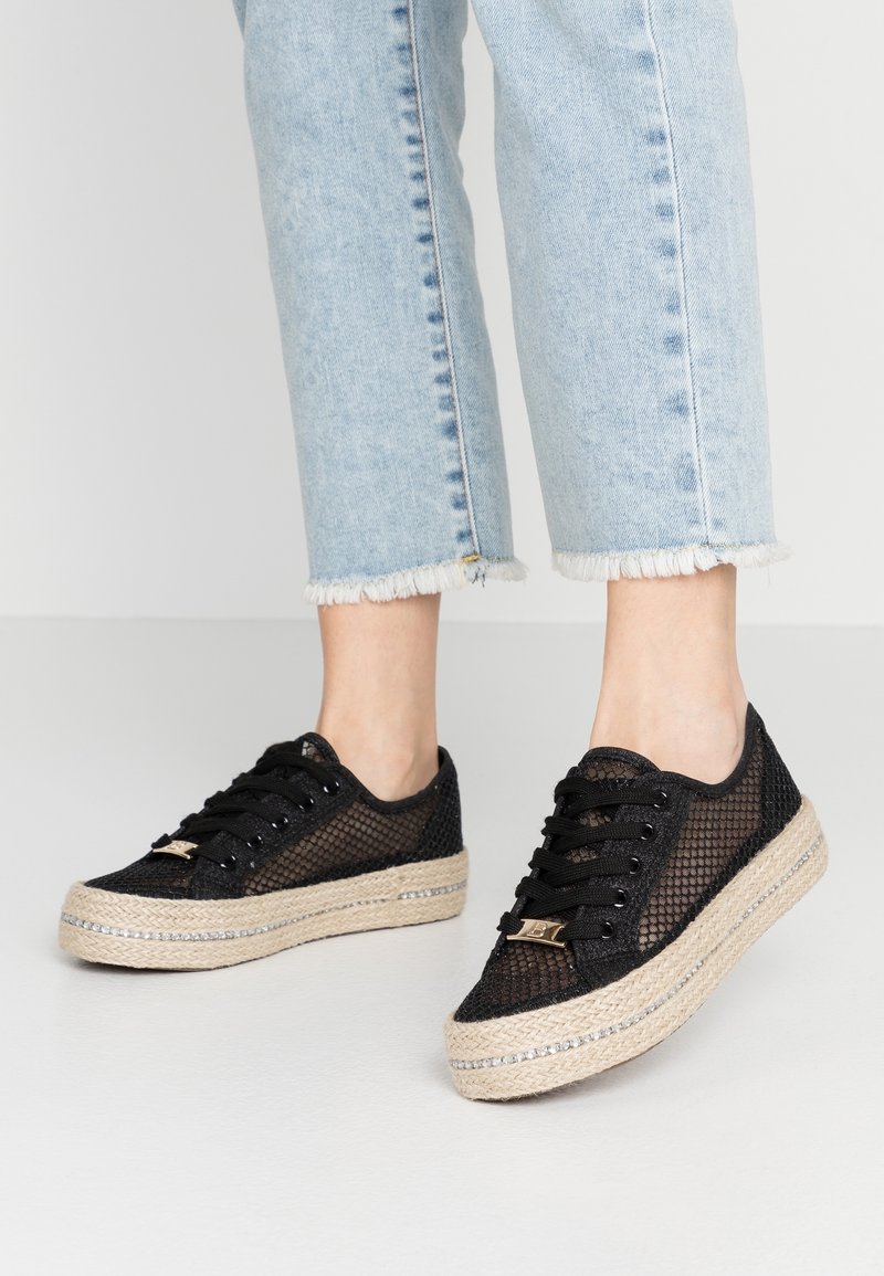 Sneaker nere in rete con una piattaforma in juta, caratterizzate da un upper testurizzato e dettagli in hardware dorato. Indossate con jeans azzurri sfilacciati.