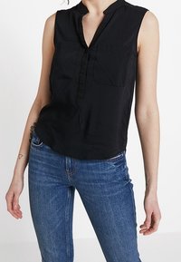 Blouse noire sans manches avec un col mao, fermeture à boutons et deux poches avant ; tissu lisse ; portée avec un jean bleu.