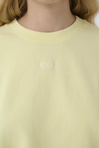 Nærbillede af person iført lysegul crewneck-shirt med lille hvidt broderet logo centreret under kraven.