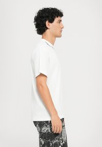 Balta trumpa rankovėmis marškinėliai su apvaliu kaklu, turintys spausdintą dizainą aplink apykaklę. Modelis stovi šoniniu profiliu prieš lengvą foną.