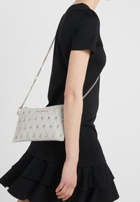 Sac à main gris clair avec un design matelassé et des clous dorés, doté d'une bandoulière chaîne. Porté à l'épaule avec une robe noire.