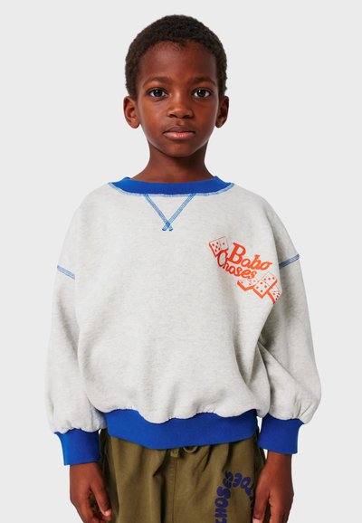 Sivi dukserica s plavim akcentima, rebrastim rukavima i donjim rubom. Na prednjoj strani se nalazi narančasti grafički prikaz s kockicama i natpisom "Baba Chases". Mekan materijal i širok kroj.