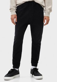 Pantalones tipo chándal negros de suave mezclilla, con cinturilla elástica, bolsillos laterales y puños estrechos, combinados con zapatillas negras.