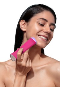 Pink handheld skincare device with a smooth texture, rounded shape, and blue light indicator, held against a model's cheek.
