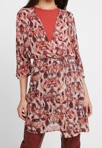 Vestido de tela fluida y ligera en rosa con patrones de paisley en rojo y marrón. Presenta un diseño envolvente, escote en forma de v y mangas de tres cuartos.