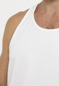 Homme portant un débardeur blanc uni sans manches, montrant l'épaule et le haut de la poitrine avec des poils visibles.