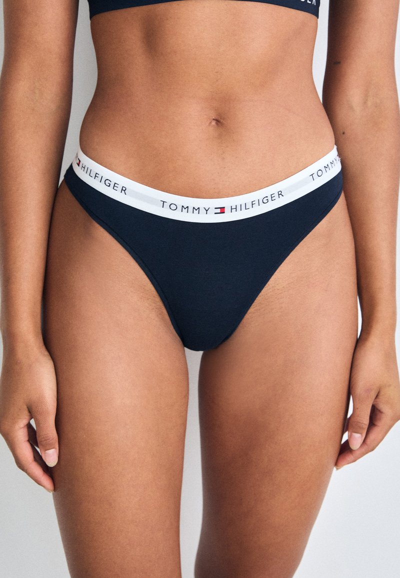 String bleu marine avec une ceinture élastique blanche arborant le logo "TOMMY HILFIGER". Le tissu est lisse et extensible.