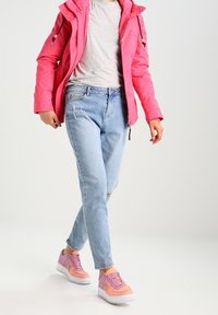 Giacca a vento rosa con cappuccio, t-shirt grigia, jeans chiari strappati e sneakers rosa con dettagli viola. Modello che cammina in avanti.