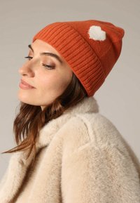 Vrouw met een roest-oranje gebreide muts met een witte hartdetail, gecombineerd met een beige nep-bontjas, die naar beneden kijkt met een lichte glimlach.