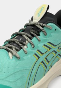 Κοντινό πλάνο από αθλητικά παπούτσια ASICS σε τυρκουάζ χρώμα, με μαύρους και άσπρους ριγέ κορδόνια και λογότυπο με κίτρινη απόχρωση στο πλάι, πάνω σε προστατευτική σόλα.