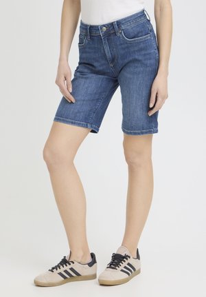 Osoba ubrana w niebieskie dżinsowe spodenki do kolan, biały top oraz beżowe sneakersy z czarnymi paskami na jednolitym tle.