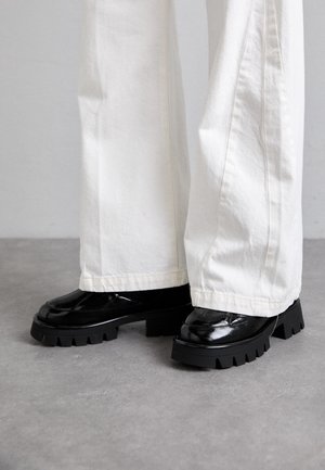 Chaussures en cuir noires à semelles épaisses portées avec un pantalon blanc long et large, debout sur un sol gris.