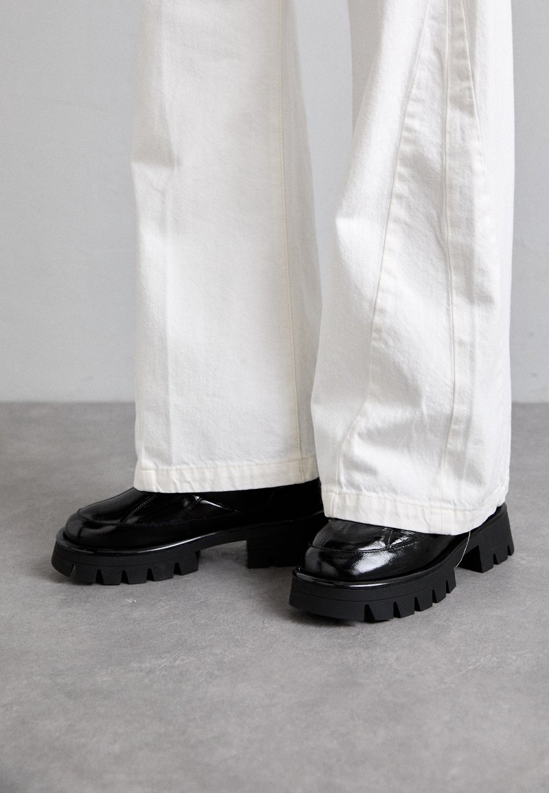 Chaussures en cuir noires à semelles épaisses portées avec un pantalon blanc long et large, debout sur un sol gris.