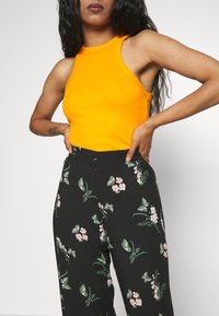 Femme portant un haut sans manches orange vif et un pantalon taille haute noir imprimé de papillons verts et de fleurs roses, les mains sur les hanches.