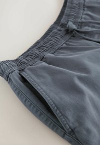 Gros plan sur un short à taille élastique bleu-gris avec poche latérale et cordon de serrage, posé sur une surface claire.